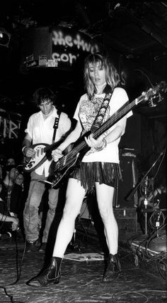 a woman playing guitar while standing on stage with other people behind her in black and white