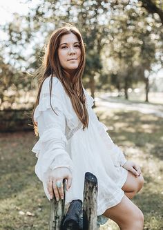 a woman in a white dress is sitting on a wooden fence and posing for the camera