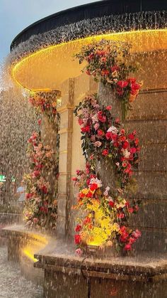 a fountain with red and pink flowers on it's sides, surrounded by rain