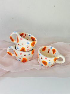 three white and orange dishes sitting on top of a pink cloth covered tablecloth next to each other