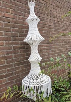 a white crocheted lamp hanging from a brick wall next to plants and bushes