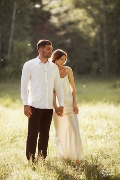 a man and woman holding hands walking through the grass in an open field with trees behind them