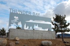 there is a sign that says thunder bay in front of some rocks and pine trees