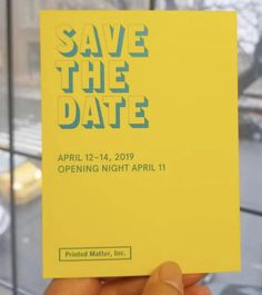 a person holding up a yellow save the date card in front of a building window