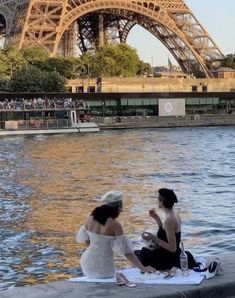 two women sitting on the edge of a body of water in front of the eiffel tower