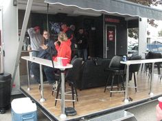 some people are standing around in front of a food truck with tables and chairs on it