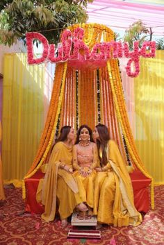 three women in yellow outfits sitting on a stage with flowers and decorations around them, posing for the camera