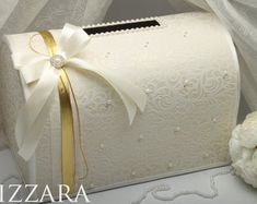 a white box with a gold ribbon tied around it and some flowers on the side