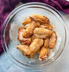 a glass bowl filled with fried chicken wings on top of a purple cloth next to a red napkin