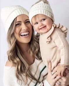 a woman holding a baby wearing a white hat and smiling at the camera with her hands on her chest