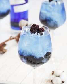 two glasses filled with blue liquid and some blackberries on top of them next to cotton floss