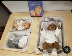 two baby dolls are sitting in small plastic trays on the table next to a book