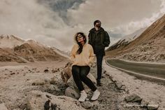 two people standing on the side of a road near some rocks and mountains with clouds in the sky