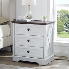 a white night stand with two drawers in front of a window and a plant next to it