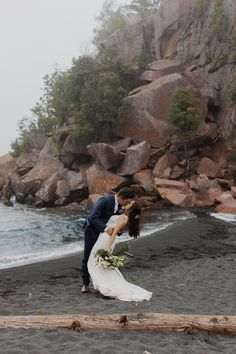 a bride and groom kissing on the beach in front of some rocks with water behind them