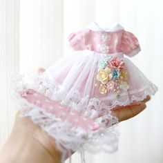 a hand holding a doll in a pink and white dress with flowers on it's chest