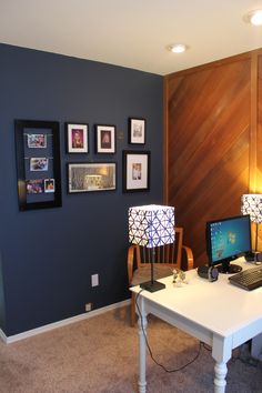 a desk with two computers on it in front of a wooden wall and framed pictures