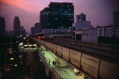 the city skyline is lit up at night with people walking on the sidewalk and cars driving down the street