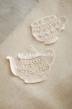 two embroidered teapots sitting on top of a piece of linen covered table cloth