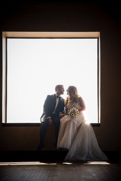 A couple sits framed by a large window; the man in a suit and the woman in a wedding dress holding a bouquet as they lean in for a kiss. Wedding Venues Indoor, Man In A Suit, Lean In, Warehouse Wedding, Large Window