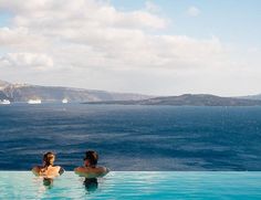 two women are sitting in the pool looking out over the water at an island and cruise ship