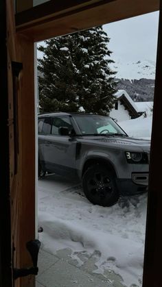 an suv is parked in the snow near a house and trees, seen through a door