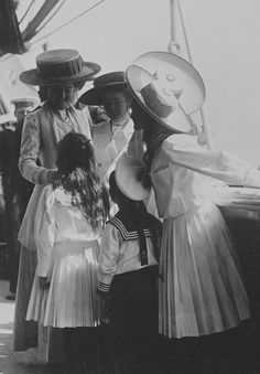 black and white photograph of three women in dresses with hats on their heads, one woman is holding the other's hand