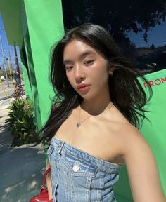 a woman with long black hair standing in front of a green wall holding a red purse