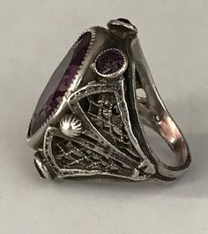 "This lovely filigreed and ornate ring has an oval shaped faceted amethyst purple color glass stone at its center. It is flanked on either side of to and bottom with small round bezel set purple glass stones. The sides have lattice shaped cut outs and the band is smooth. The face of this ring is about 3/4\" long and about 1/2\" across. It is a size 3 1/4 and is marked Sterling. Please note there are some overall signs of wear, especially to the stones, four of five of which, including the center Heirloom Purple Amethyst Ring With Intricate Design, Unique Oval Filigree Ring For Anniversary, Vintage Silver Amethyst Ring With Accent Stones, Heirloom Oval Filigree Ring, Heirloom Style Purple Amethyst Ring With Filigree, Unique Oval Amethyst Ring For Formal Occasions, Vintage Amethyst Ring With Stone Setting, Oval Filigree Ring With Stone Setting For Formal Occasions, Vintage Oval Ruby Ring With Accent Stones