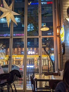 people sitting at tables in front of a restaurant window with lights on the outside and inside