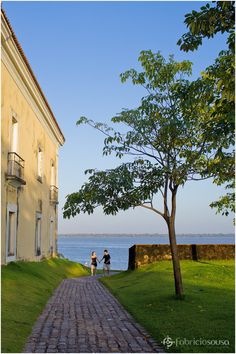 two people walking down a brick path next to a building near the water on a sunny day