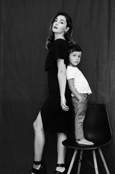 a woman standing next to a little boy on top of a chair in front of a black backdrop