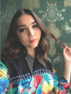 a woman with long hair wearing a colorful shirt and pearls on her collar, posing in front of a floral wallpaper