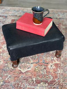 a coffee cup sitting on top of a black ottoman next to a red and white book