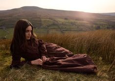 a woman laying on the ground in a field with hills and grass behind her, wearing a brown dress