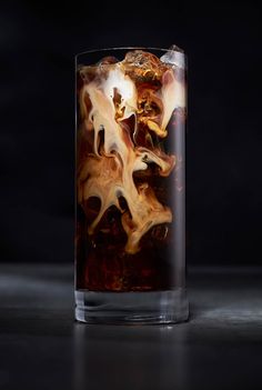 a glass filled with ice and liquid on top of a black table next to a dark background