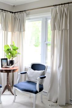 a living room with two chairs and a table in front of a window that has white drapes on the windowsill