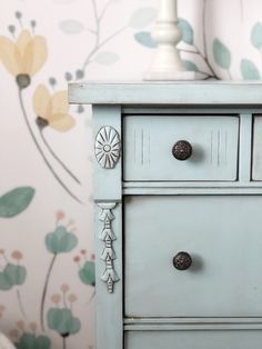 a blue dresser with drawers and knobs in front of a floral wallpapered wall