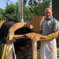 a man in an apron is holding a tray with food on it while standing next to a brick oven