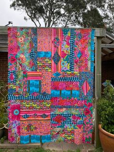 a colorful quilt hanging on the side of a building next to a potted plant