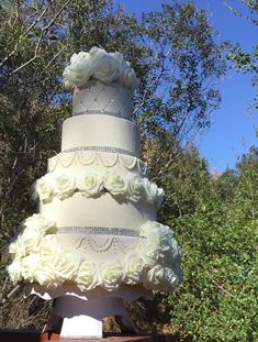 a three tiered cake with white flowers on the top is sitting in front of some trees