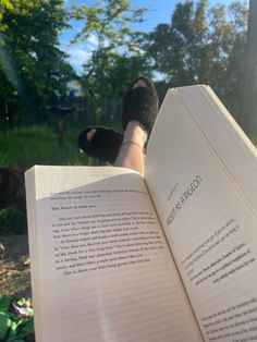 an open book sitting on top of a person's feet in the grass next to trees