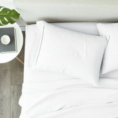 a bed with white sheets and pillows on top of it next to a table with a plant