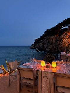 an outdoor dining set up on the beach at night with lit candles in front of it