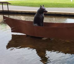 a statue of a dog sitting on top of a boat in the water next to a park