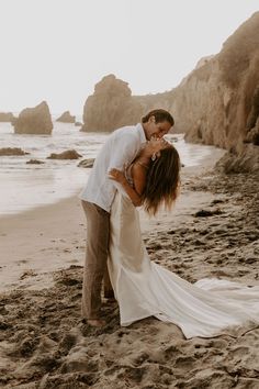 a bride and groom kissing on the beach in front of some rocky cliffs at sunset