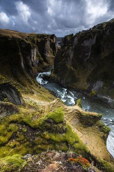 a river flowing through a lush green valley next to a rocky cliff side under a cloudy sky