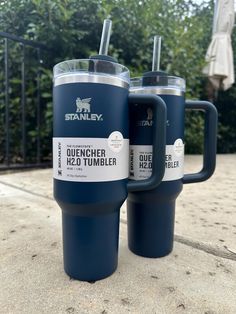 two blue travel mugs sitting next to each other on the ground near some bushes