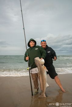 two people standing on the beach with a fish and fishing pole in front of them