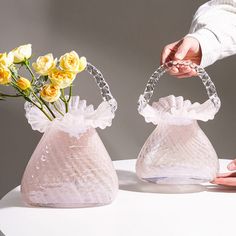 two clear vases with yellow flowers in them on a white table and one is holding a hand